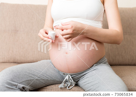 a pregnant girl sits at home on the bed and smears smile an anti-stretch mark cream on her stomach. Pregnancy, motherhood, preparation and expectation concept 73065834
