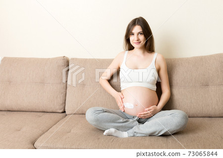 a pregnant girl sits at home on the bed and smears an anti-stretch mark cream on her stomach. Pregnancy, motherhood, preparation and expectation concept 73065844