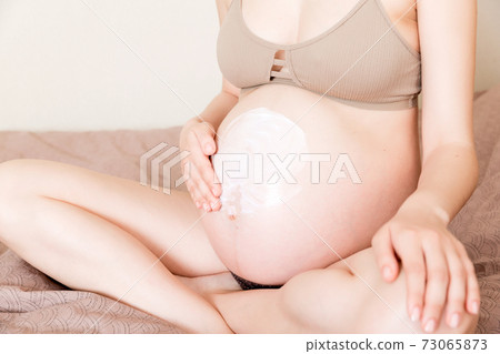 a pregnant girl sits at home on the bed and smears an anti-stretch mark cream on her stomach. Pregnancy, motherhood, preparation and expectation concept a pregnant girl sits at home on the bed and smears an anti-stretch mark cream on her stomach. Pregnancy, motherhood, preparation and expectation concept 73065873