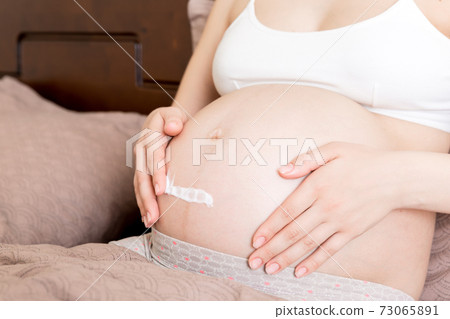 a pregnant girl sits at home on the bed and smears an anti-stretch mark cream on her stomach. Pregnancy, motherhood, preparation and expectation concept a pregnant girl sits at home on the bed and smears an anti-stretch mark cream on her stomach. Pregnancy, motherhood, preparation and expectation concept 73065891