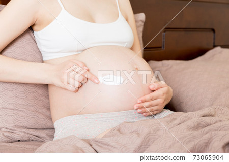 a pregnant girl sits at home on the bed and smears an anti-stretch mark cream on her stomach. Pregnancy, motherhood, preparation and expectation concept a pregnant girl sits at home on the bed and smears an anti-stretch mark cream on her stomach. Pregnancy, motherhood, preparation and expectation concept 73065904