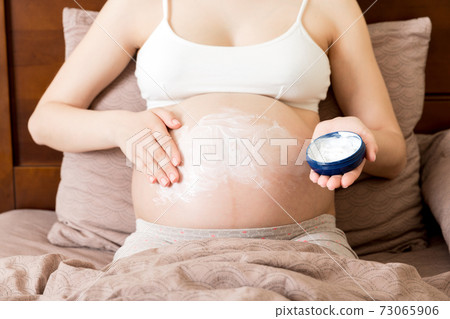 a pregnant girl sits at home on the bed and smears smile an anti-stretch mark cream on her stomach. Pregnancy, motherhood, preparation and expectation concept 73065906