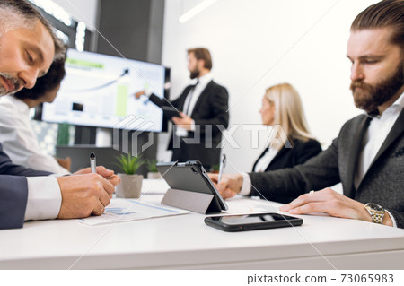 Two focused diverse businessmen, making notes while listening to the speech of male speaker, showing financial presentation to diverse employees group at office meeting. Focus on tablet and phone Two focused diverse businessmen, making notes while listening to the speech of male speaker, showing financial presentation to diverse employees group at office meeting. Focus on tablet and phone 73065983