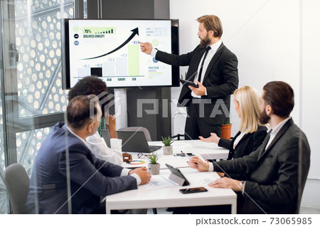 Front view of young confident bearded businessman, showing statistic datas on big plasma screen for his focused high-skilled multiethnical colleagues during meeting in office room Front view of young confident bearded businessman, showing statistic datas on big plasma screen for his focused high-skilled multiethnical colleagues during meeting in office room 73065985