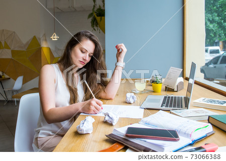 Concentrated young woman making notes on paper while working on her laptop at a cafe, coworking. Brainstorm process. Remote work concept. Freelancer workspace. Copy space. 73066388