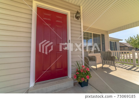 Shiny red wooden front door of a home with wicker chairs on the sunlit porch Shiny red wooden front door of a home with wicker chairs on the sunlit porch 73069185
