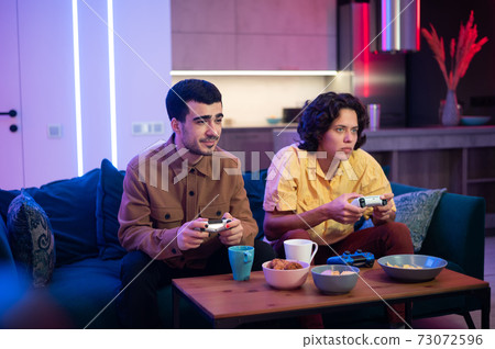 Happy male gamers playing video games at home. Guy with manicure holding gaming console joystick. 73072596