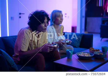 Couple of young adults playing video games at home. Emotional diverse gamers holding joysticks and compete in intense video game on gaming console. 73072604