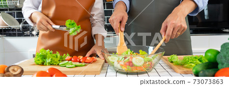 Happy couple having fun standing in kitchen at home preparing vegetable salad husband and wife vegetarians chop vegetables prepare for dinner in loft kitchen at home. 73073863