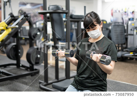 A woman wearing a mask and doing muscle training at a fitness gym A woman wearing a mask and doing muscle training at a fitness gym 73087950