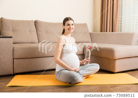 Pregnant young woman with bottle of water while sitting in lotus position on yoga mat. Maintaining water balance at coronavirus time 73089511