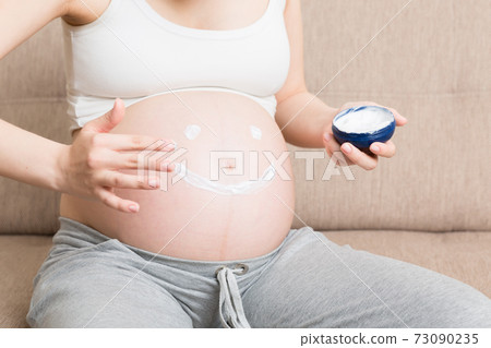 a pregnant girl sits at home on the bed and smears smile an anti-stretch mark cream on her stomach. Pregnancy, motherhood, preparation and expectation concept a pregnant girl sits at home on the bed and smears smile an anti-stretch mark cream on her stomach. Pregnancy, motherhood, preparation and expectation concept 73090235