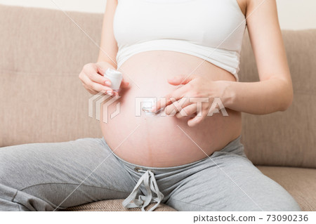 a pregnant girl sits at home on the bed and smears smile an anti-stretch mark cream on her stomach. Pregnancy, motherhood, preparation and expectation concept a pregnant girl sits at home on the bed and smears smile an anti-stretch mark cream on her stomach. Pregnancy, motherhood, preparation and expectation concept 73090236