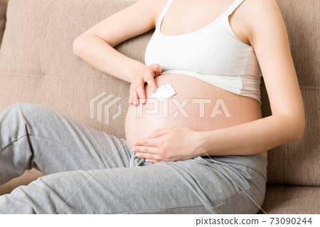 a pregnant girl sits at home on the bed and smears an anti-stretch mark cream on her stomach. Pregnancy, motherhood, preparation and expectation concept a pregnant girl sits at home on the bed and smears an anti-stretch mark cream on her stomach. Pregnancy, motherhood, preparation and expectation concept 73090244
