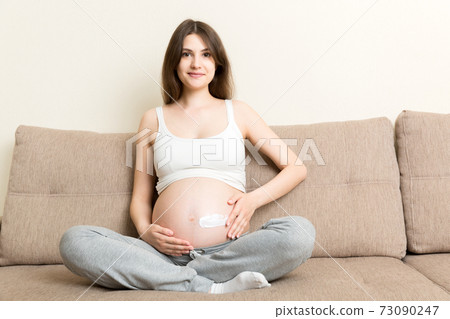 a pregnant girl sits at home on the bed and smears an anti-stretch mark cream on her stomach. Pregnancy, motherhood, preparation and expectation concept a pregnant girl sits at home on the bed and smears an anti-stretch mark cream on her stomach. Pregnancy, motherhood, preparation and expectation concept 73090247