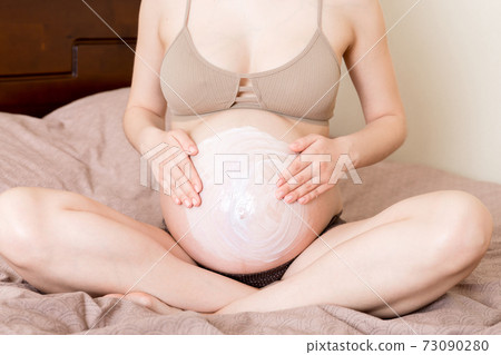 a pregnant girl sits at home on the bed and smears an anti-stretch mark cream on her stomach. Pregnancy, motherhood, preparation and expectation concept 73090280