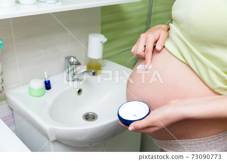 Young pregnant woman applying anti-stretch marks cream to the abdomen in the morning in the bathroom. Pregnancy, maternity and bodycare Young pregnant woman applying anti-stretch marks cream to the abdomen in the morning in the bathroom. Pregnancy, maternity and bodycare 73090773