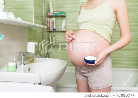 Young pregnant woman applying anti-stretch marks cream to the abdomen in the morning in the bathroom. Pregnancy, maternity and bodycare Young pregnant woman applying anti-stretch marks cream to the abdomen in the morning in the bathroom. Pregnancy, maternity and bodycare 73090774