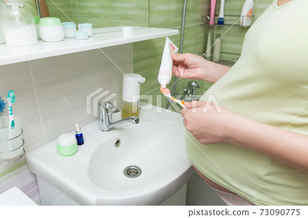 Young pregnant woman dressed in a robe in the bathroom at home. Cute adult girl in the third trimester of pregnancy in the home bathroom brushing her teeth 73090775