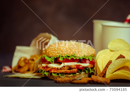 Burger on a rustic wooden board and unhealthy snacks and chips Burger on a rustic wooden board and unhealthy snacks and chips 73094185