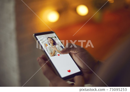 Man holding cellphone, looking at young woman's profile picture on dating app and pressing like 73095253