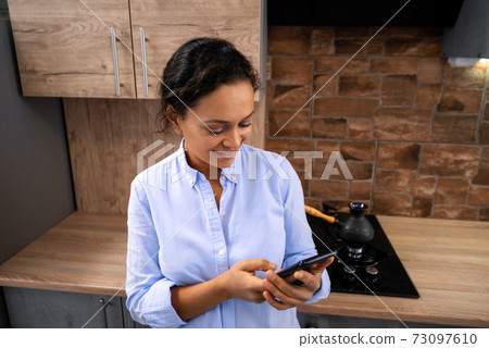 Young woman writes a message on the smartphone while standing in the kitchen. 73097610