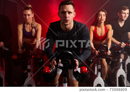 muscular young caucasian man exercising cardio training on bicycle in fitness gym with friends muscular young caucasian man exercising cardio training on bicycle in fitness gym with friends 73098809