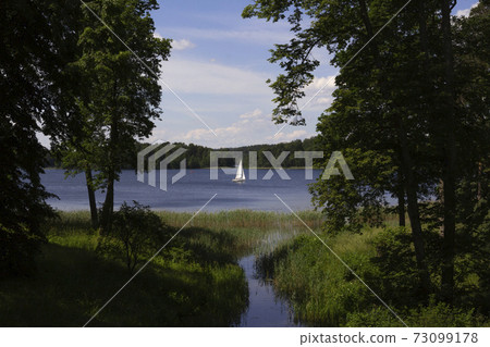 Beautiful landscape - green trees and meadows, a white boat sails in the distance of the lake. Lithuania, near Trakai Beautiful landscape - green trees and meadows, a white boat sails in the distance of the lake. Lithuania, near Trakai 73099178