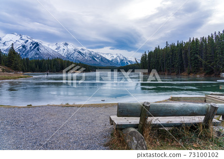 Johnson Lake frozen water surface in winter time. Snow-covered mountain in the background. Tourists here doing ice-skating in this season. Banff National Park, Canadian Rockies, Alberta, Canada. 73100102