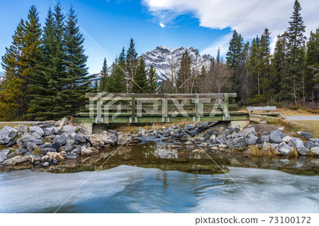Wooden footbridge in the forest across frozen icy water surface in winter. Snow-covered mountain in background. Johnson lake, Banff National Park, Canadian Rockies, Alberta, Canada. Wooden footbridge in the forest across frozen icy water surface in winter. Snow-covered mountain in background. Johnson lake, Banff National Park, Canadian Rockies, Alberta, Canada. 73100172