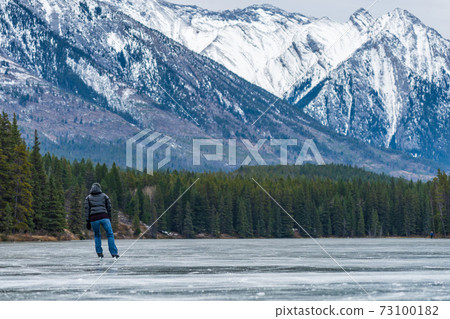 Tourists doing ice-skating in Johnson Lake frozen water surface in winter time. Snow-covered mountain in the background. Banff National Park, Canadian Rockies, Alberta, Canada. 73100182