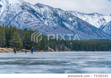 Tourists doing ice-skating in Johnson Lake frozen water surface in winter time. Snow-covered mountain in the background. Banff National Park, Canadian Rockies, Alberta, Canada. Tourists doing ice-skating in Johnson Lake frozen water surface in winter time. Snow-covered mountain in the background. Banff National Park, Canadian Rockies, Alberta, Canada. 73100183