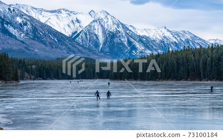 Tourists doing ice-skating in Johnson Lake frozen water surface in winter time. Snow-covered mountain in the background. Banff National Park, Canadian Rockies, Alberta, Canada. Tourists doing ice-skating in Johnson Lake frozen water surface in winter time. Snow-covered mountain in the background. Banff National Park, Canadian Rockies, Alberta, Canada. 73100184