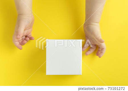 Children's hands and white cardboard box on yellow background, top view. Copy space. Mock up. 73101272