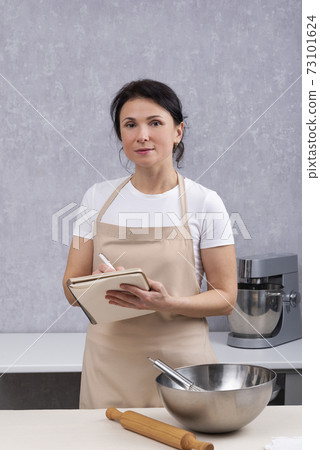 Portrait of woman chef in kitchen with cookbook in her hands next to bowl and rolling pin. Vertical frame 73101624