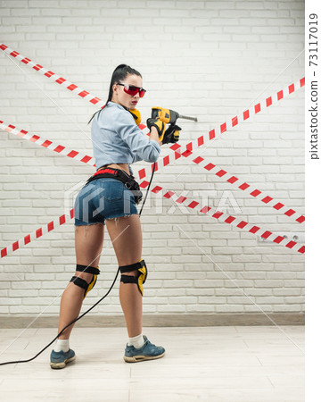 a slender woman in the image of a construction worker with a puncher against a white textured brick wall a slender woman in the image of a construction worker with a puncher against a white textured brick wall 73117019
