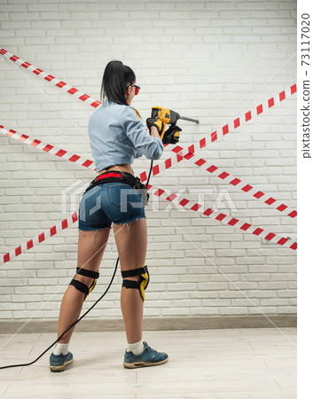 a slender woman in the image of a construction worker with a puncher against a white textured brick wall a slender woman in the image of a construction worker with a puncher against a white textured brick wall 73117020