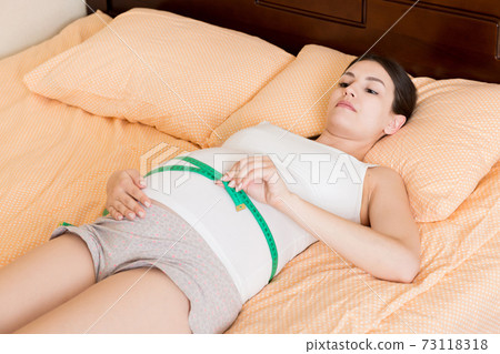 Young beautiful pregnant girl measuring her belly with a tape in home on bed. Healthy pregnancy concept 73118318