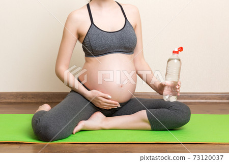 Pregnant young woman with bottle of water while sitting in lotus position on yoga mat. Maintaining water balance at coronavirus time 73120037