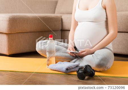 Pregnant female holding smartphone while sitting on exercise yoga mat at home at coronavirus time Pregnant female holding smartphone while sitting on exercise yoga mat at home at coronavirus time 73120096