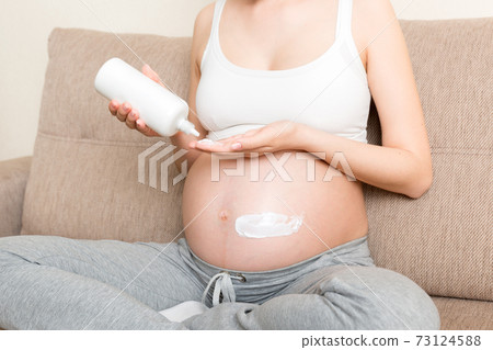 a pregnant girl sits at home on the bed and smears smile an anti-stretch mark cream on her stomach. Pregnancy, motherhood, preparation and expectation concept a pregnant girl sits at home on the bed and smears smile an anti-stretch mark cream on her stomach. Pregnancy, motherhood, preparation and expectation concept 73124588