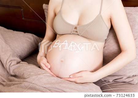 a pregnant girl sits at home on the bed and smears an anti-stretch mark cream on her stomach. Pregnancy, motherhood, preparation and expectation concept. the word baby on the belly a pregnant girl sits at home on the bed and smears an anti-stretch mark cream on her stomach. Pregnancy, motherhood, preparation and expectation concept. the word baby on the belly 73124633