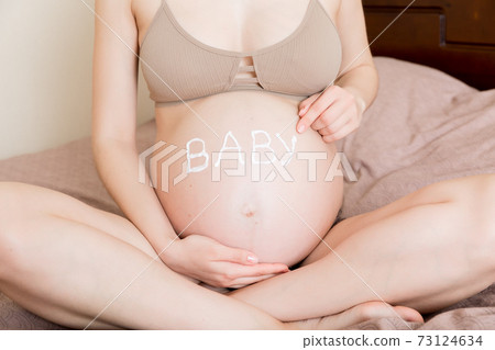 a pregnant girl sits at home on the bed and smears an anti-stretch mark cream on her stomach. Pregnancy, motherhood, preparation and expectation concept. the word baby on the belly 73124634