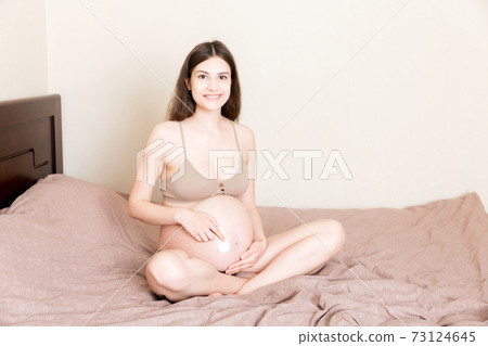 a pregnant girl sits at home on the bed and smears an anti-stretch mark cream on her stomach. Pregnancy, motherhood, preparation and expectation concept a pregnant girl sits at home on the bed and smears an anti-stretch mark cream on her stomach. Pregnancy, motherhood, preparation and expectation concept 73124645