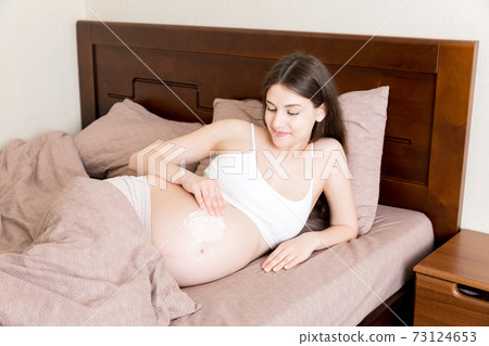 a pregnant girl sits at home on the bed and smears an anti-stretch mark cream on her stomach. Pregnancy, motherhood, preparation and expectation concept 73124653