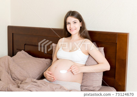a pregnant girl sits at home on the bed and smears an anti-stretch mark cream on her stomach. Pregnancy, motherhood, preparation and expectation concept a pregnant girl sits at home on the bed and smears an anti-stretch mark cream on her stomach. Pregnancy, motherhood, preparation and expectation concept 73124670