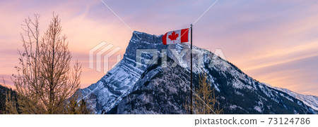 Close up of National Flag of Canada. Mount Rundle and dried trees in the background. Banff National Park, Canadian Rockies, Canada. 73124786