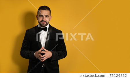 Handsome man in tuxedo standing fingers folded down smiling on camera, work profession lifestyle. Handsome young smiling caucasian man isolated on yellow background 73124832