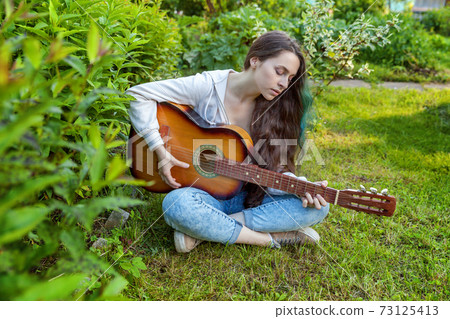 Young woman sitting in grass and playing guitar 73125413