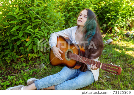 Young woman sitting in grass and playing guitar 73125509